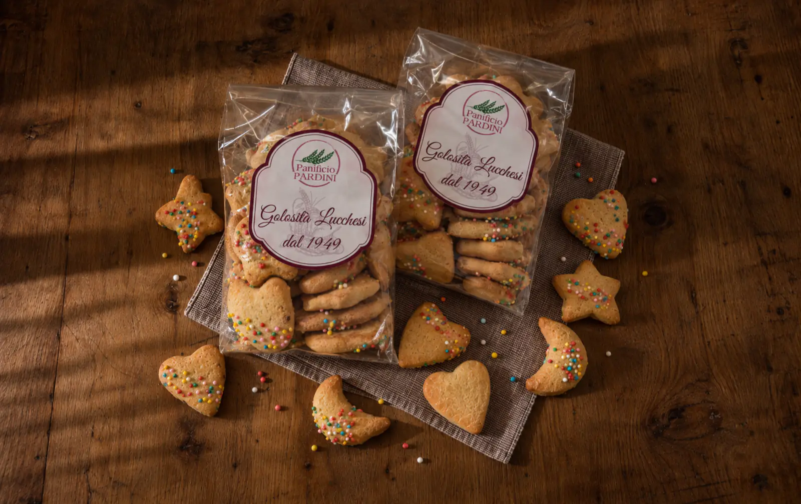 Due sacchetti di befanini del Panificio Pardini e biscotti sfusi a forma di cuore, stella e luna con i chicchi colorati, su un tavolo di legno.