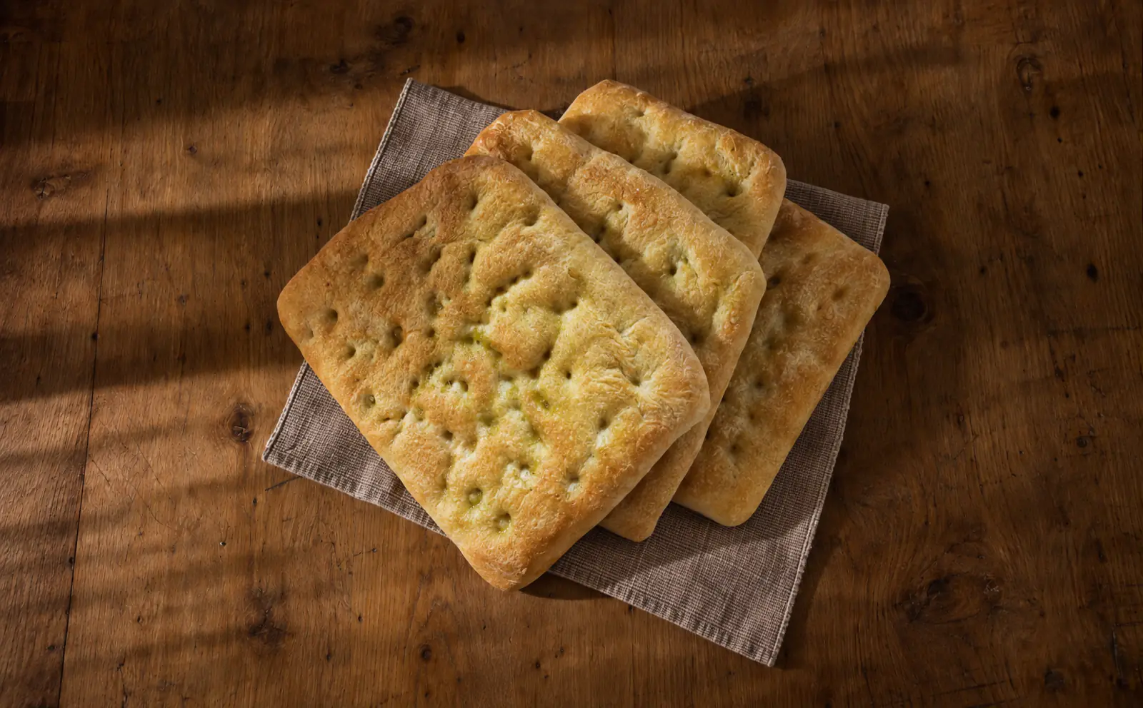 Le nostre focacce dorate all’olio, con i classici buchi, appoggiate su un tovagliolo su un tavolo di legno.