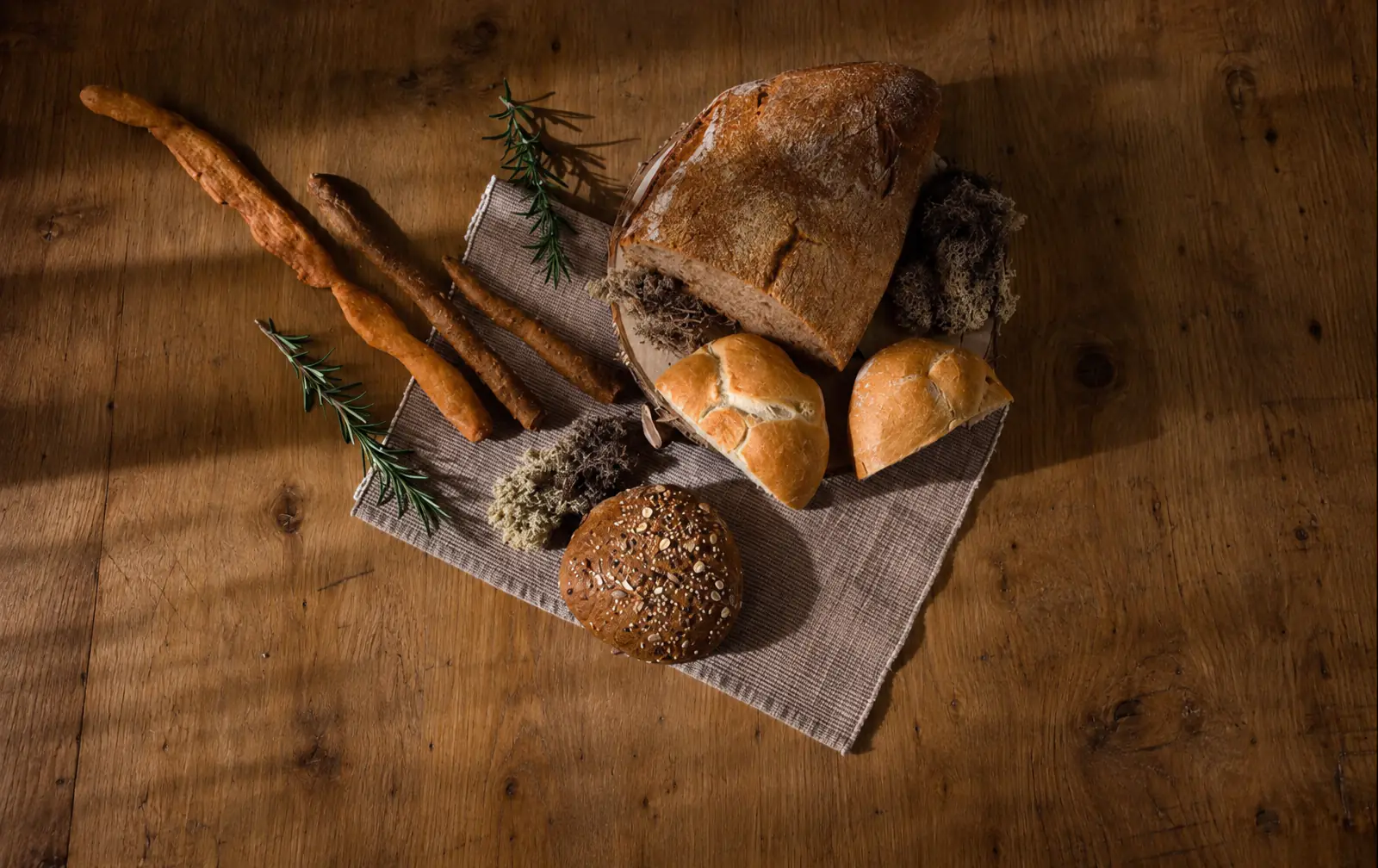 Il nostro pane su un tavolo di legno: un filone infarinato, un panino tagliato a metà, una pagnotta ai semi e grissini, con rametti di rosmarino.